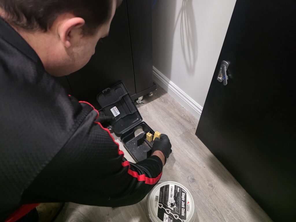East West Pest technician servicing a rodent station beside a cabinet
